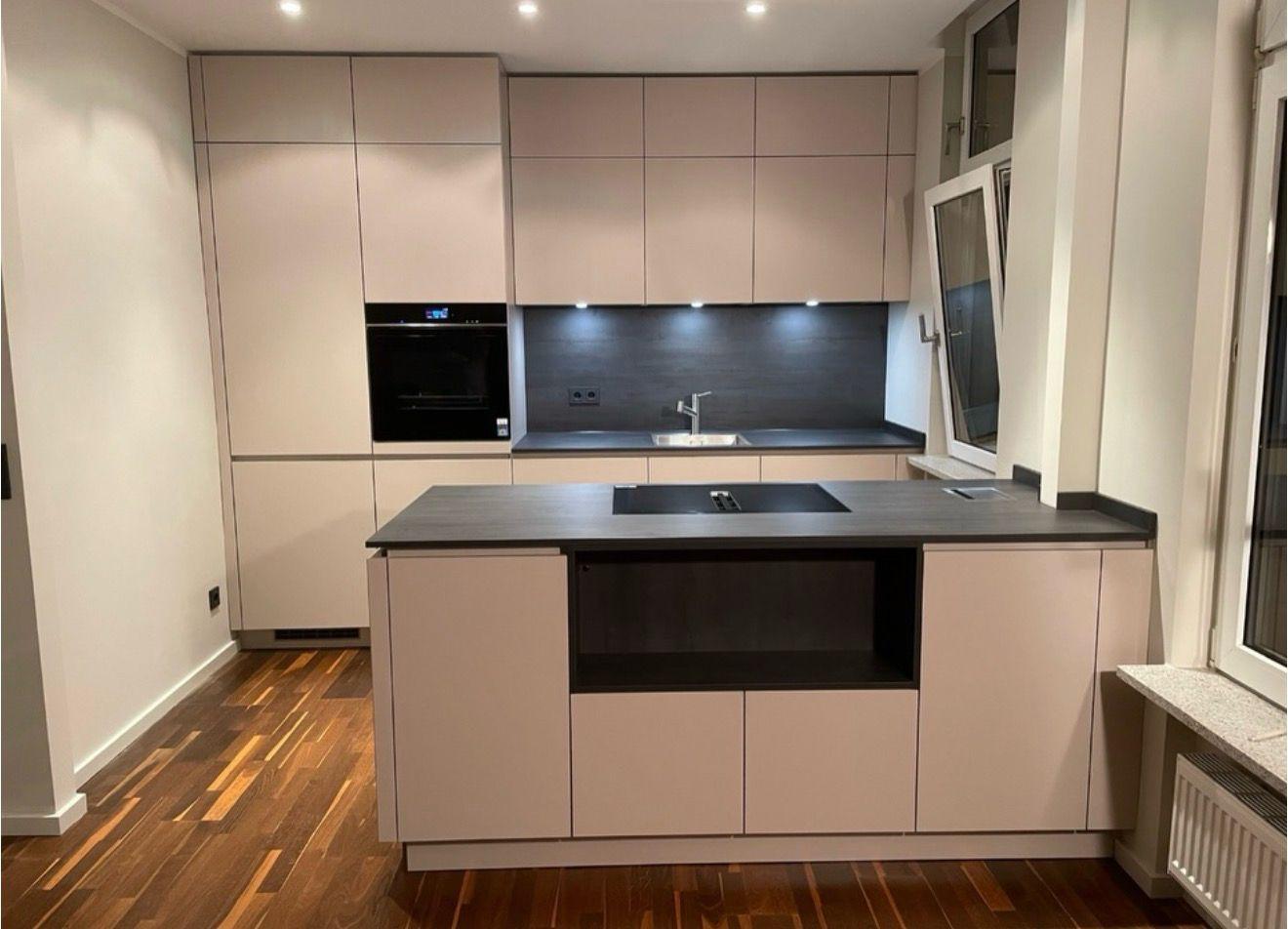 Moderne, grifflose Einbauküche in Taupe/Grau mit freistehender Kochinsel, dunkler Arbeitsplatte, integriertem Backofen und Holzboden.