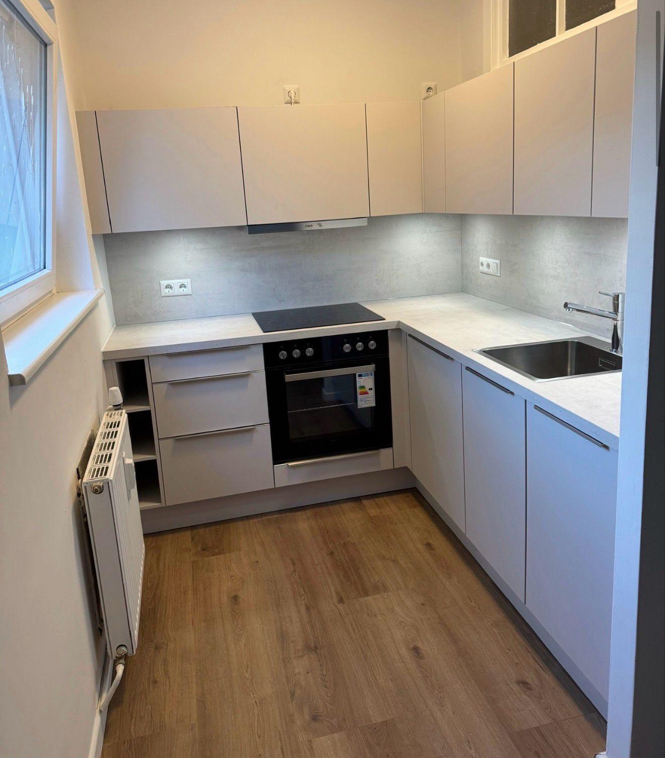 Kleine, helle L-Küche in Hellgrau mit integriertem Backofen, Induktionskochfeld und Spülbecken aus Edelstahl unter Oberschränken.