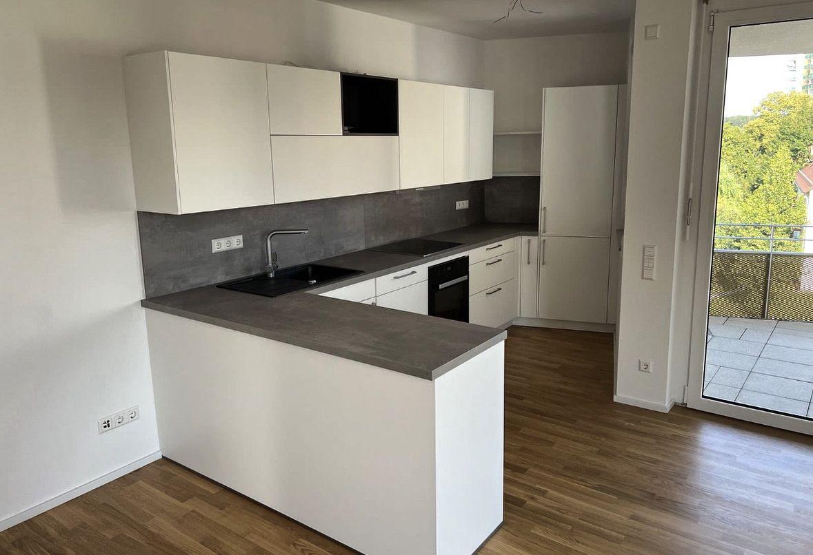 Offene L-förmige Küche in Weiß mit grauer Arbeitsplatte, schwarzen Küchengeräten und Zugang zum Balkon.